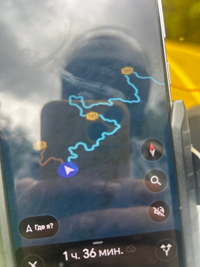 This image shows a smartphone screen displaying a navigation application. The application shows a map with a blue line indicating a route and several yellow markers with the number "185" on them, suggesting road numbers or points of interest. A blue arrow symbol indicates the current location or direction of travel. There are also several circular icons on the screen, including a compass, a magnifying glass for search, a symbol that may indicate a mute function, and a symbol representing a junction or turn. At the bottom of the screen, text displays "1 ч. 36 мин.", indicating a duration of 1 hour and 36 minutes. Below this, there is a button with Russian text "Где я?", which translates to "Where am I?". The overall environment appears to be a vehicle, as suggested by the reflections on the screen, the angle of the phone, and the yellow background visible through the phone's screen which could be a road or surrounding landscape. There is no discernible information within the image that specifically places it in Gopeng, Malaysia.