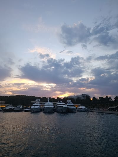The image depicts a harbor scene at dusk in Vouliagmeni Municipal Unit, Greece. Several large yachts are moored in the foreground, with their sleek, modern designs illuminated by the fading light. The water in the harbor is calm, reflecting the twilight sky. In the background, a lush green treeline borders the water, with a hotel or large building visible nestled within the trees. The sky is a canvas of soft purples, oranges, and grays, with scattered clouds suggesting the transition from day to night. The overall atmosphere is serene and picturesque.
