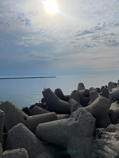 The image depicts a seascape with a breakwater constructed of concrete tetrapods in the foreground. The tetrapods are angular, grey, and appear weathered, stacked closely together. Beyond the breakwater, a calm, blue sea extends to the horizon, where a long, low line of breakwater structures forms a barrier, possibly a harbor entrance. In the distance, a small lighthouse or navigational marker is visible on this distant breakwater, and a few boats, including a larger ship, are present on the water. The sky above is partly cloudy, with the sun shining through, creating a bright area in the upper right quadrant of the image. The scene is likely located in Baltiysk, Russia, given the context of a Baltic Sea port.