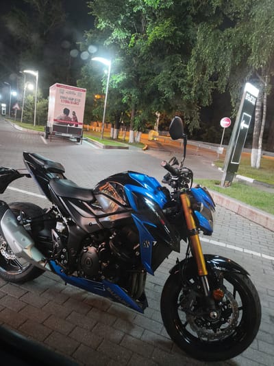 A blue and black Suzuki motorcycle is parked on a brick-patterned pavement at night. The motorcycle is positioned at an angle, with its front wheel slightly turned. In the background, a white trailer with a large advertisement is visible. The advertisement features text in Russian and an image of two people. Streetlights illuminate the scene, and trees with dense foliage line the background. A black sign with white lettering, also in Russian, stands to the right of the motorcycle, with an arrow pointing to the left. A red and white circular traffic sign is also present. The overall environment appears to be an outdoor public space, possibly a parking lot or a roadside area.
