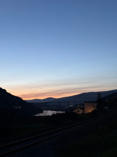 The image depicts a twilight scene in Peso da Régua, Portugal, looking towards the Douro River valley. The upper two-thirds of the frame features a clear sky transitioning from a deep blue overhead to a band of light blue, then a faint orange and pink hue at the horizon. The lower third is dominated by terrestrial features rendered largely in silhouette due to low light conditions.

In the foreground, two parallel railway tracks extend from the lower left, curving gently to the right as they recede into the mid-distance. The tracks consist of dark metal rails laid on a bed of dark ballast. Dark, indistinct vegetation borders the railway line.

The midground features a body of water, likely the Douro River, appearing as a silvery-white strip reflecting ambient light, horizontally spanning a portion of the scene. On the right side of the riverbank, a building with light-colored walls glows with internal or nearby artificial illumination, providing a yellowish contrast against the dim environment. A tall, dark coniferous tree stands adjacent to this illuminated structure.

In the background, successive layers of dark hills and mountains are silhouetted against the horizon. A long, multi-span bridge crosses the valley between these hills, its structure punctuated by numerous small, distant lights, indicating active roadways. Further up on the distant hillsides, scattered clusters of small, bright lights denote urban or residential areas. On the far right horizon, faint, tall structures resembling wind turbines are visible against the sky. No individuals or specific actions are discernible within the scene.