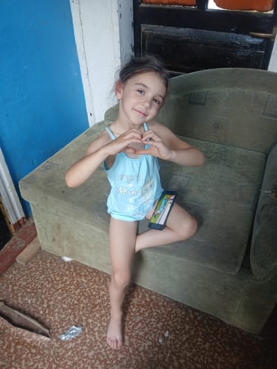 A young girl is lying on a green sofa, holding a smartphone in her lap. She is wearing a light blue tank top with a graphic of Elsa from Frozen and the words "Sparkle like Magic" printed on it. Her left arm is bent, with her fingers near her mouth, and her right leg is extended. The sofa appears worn, with a faded pattern. Beside the sofa, on the floor, is a piece of crumpled aluminum foil. The background shows a blue wall and a dark, window-like structure with orange panes. The scene suggests an indoor setting, possibly a living room. The lighting appears to be natural, indicating it might be daytime. The girl's expression is relaxed, perhaps indicating she is engaged with the phone. The location context is Pishchanka, Ukraine.
