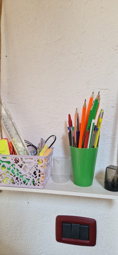 The image shows a close-up of a white, textured wall with a white shelf attached. On the shelf, there are several items, including a light purple basket filled with colorful stationery such as highlighters and pens. To the right of the basket, a green cup holds various pens and pencils. Further to the right, a small clear cup and a pencil sharpener are visible. Below the shelf, a dark red light switch with three black buttons is mounted on the wall. The overall scene appears to be an indoor setting, possibly a desk or study area. The image does not contain any specific landmarks or visual cues that would definitively place it in Brusson, Italy.