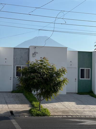 This is an outdoor, eye-level, vertical shot of a residential street in San Miguel, El Salvador. The foreground shows a grey asphalt road with a white dashed line and a storm drain. Adjacent to the road is a tiled driveway with a narrow strip of green grass running alongside it. A young tree with green foliage is situated in the grass strip. In the background are two identical, modern houses, numbered 1 and 2. House 1 has a white front door, a grey textured accent wall, and a white upper facade. House 2 has a white front door, a green textured accent wall, and a white upper facade. Both houses have white framed windows and white upper walls. Electrical wires are visible strung across the clear blue sky, with a faint, pale mountain range in the far distance. The sky is bright and clear, suggesting daytime.