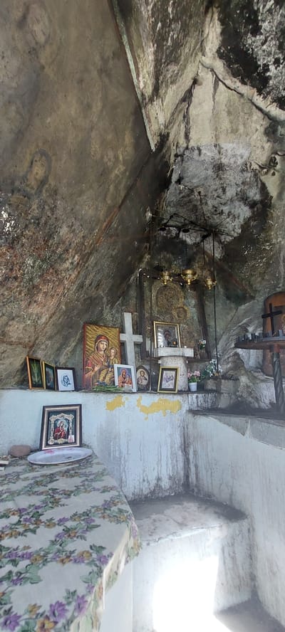 This image shows the interior of a room or alcove in Бачково, Bulgaria. The walls and ceiling appear to be made of rough stone, and there are several religious icons and crosses mounted on the walls. A chandelier hangs from the ceiling. The room is small and dimly lit. A patterned fabric drapes over a surface on the right side of the frame.