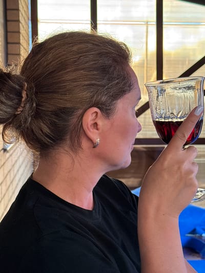 A close-up, vertically oriented shot shows a person drinking from a glass of red liquid, possibly wine. The person's head is tilted back, and their lips are touching the rim of the glass, which is held by another person's hand. The hand has a light gray fingernail polish. The person drinking is wearing a black t-shirt and has their hair pulled back in a bun. They are also wearing an earring with a delicate, almost triangular design. The background appears to be outdoors, with some sort of fence or railing and diffused light suggesting daytime. The overall mood is relaxed and possibly celebratory.
