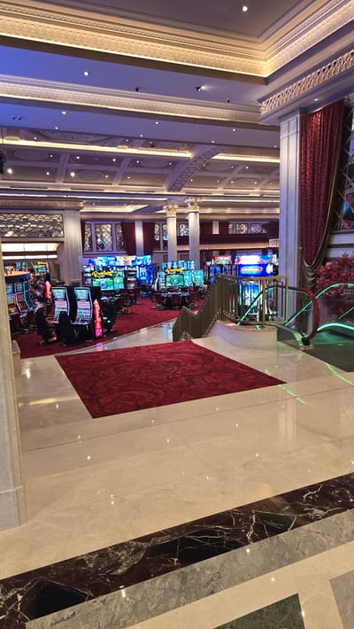 The image depicts the interior of a luxurious casino. The foreground features a polished marble floor with decorative dark and light stone inlays. A broad red carpet extends from the bottom of the frame towards the casino floor. To the right, a modern escalator with green illuminated accents ascends. In the midground, the casino floor is filled with numerous brightly lit slot machines, some with people seated and playing. Red carpets are visible throughout the gaming area. The background reveals the elaborate architecture of the casino, including ornate ceilings with detailed moldings and recessed lighting, as well as large columns and red velvet curtains. The overall impression is one of opulence and entertainment. The image is not specific to Agios Epiktitos, Northern Cyprus / Cyprus, as it depicts a generic casino interior that could be located anywhere in the world.