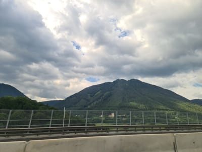 The image depicts a scenic view of a mountainous landscape with a cloudy sky, captured from a moving vehicle. In the foreground, a concrete barrier and a metal fence are visible, indicating the viewpoint is from a highway or elevated road. Beyond the fence, a green, forested mountain dominates the scene, with a building complex visible on its slope. The sky is filled with a mix of dark and light gray clouds, with patches of blue sky peeking through. The overall impression is one of a vast natural landscape under a dynamic sky. The location is identified as Schottwien, Austria, a town known for its mountainous terrain and picturesque views.