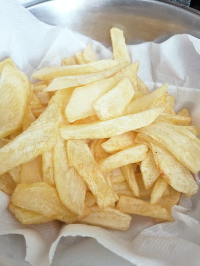 This is a close-up, overhead shot of a pile of golden-brown french fries. They are resting on a layer of white paper towels, which are partially visible at the bottom and sides of the frame. The fries are cut into thick, irregular strips, and many of them have a slightly glistening appearance, suggesting they have recently been fried. Some of the fries show a hint of reddish-brown on their edges, indicating they are well-cooked. The background shows the edge of a metal bowl, likely where the fries are being drained. The lighting is bright, suggesting it is daytime, and there are no obvious location cues beyond the implied setting of a kitchen or food preparation area. The image is entirely focused on the food, with no people or other elements visible.