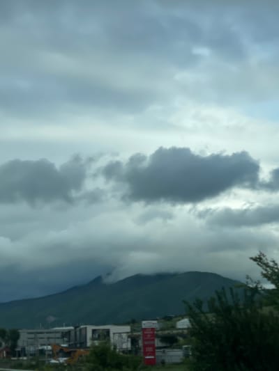 The image depicts a cloudy sky over Gorovani, Georgia. A mountain range is visible on the right side of the frame, next to a small settlement consisting of buildings, a sign, and a construction excavator. Dominant heavy clouds obscure the sky.