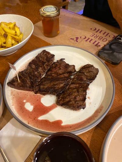 The image shows a plate of grilled steak with a red sauce on a wooden table. The steak is cooked to medium-rare and has grill marks on it. There is a glass of red wine next to the plate. The plate is white with a gray rim and is slightly angled towards the right of the image. The steak is arranged in a circular pattern, with the thickest piece in the middle. There is a small flag with a red "1" on it stuck into the steak. The image is likely taken at a restaurant in Punta Hermosa, Peru, as this is a popular tourist destination. The image was taken in the afternoon and is from a close-up angle, with the focus on the food. The image appears to be taken on a mobile phone. The restaurant is decorated with a wooden table and a brown placemat with the restaurant name on it. The background includes a small jar of honey and a cell phone in a case with an abstract design, all in a blurred background. There is a partial view of a person's arm on the top right of the image, as well as the edge of a wooden chair. There is a small plate on the lower right of the image that is out of focus.