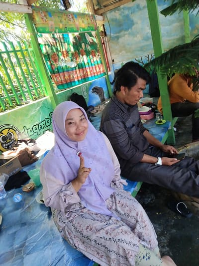 The image shows a woman in a light purple hijab and floral print pants, holding up her index finger which has red ink on it, indicating she has voted. She is smiling at the camera. To her right, a man is sitting and looking at his phone. Behind them, another man is sitting and appears to be looking at something. They are sitting on a blue painted bench, in what seems to be an outdoor sitting area. There is a mural of a sky and other cartoonish paintings on the walls. The scene is located in Palohseulimeng, Indonesia. There are cups, plates, and other items suggesting they have been eating.