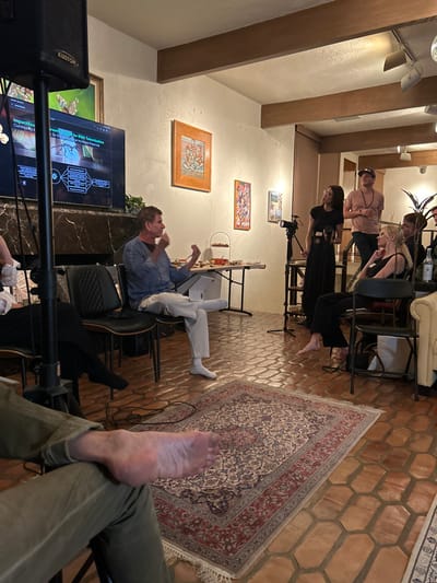 The image depicts an indoor gathering, likely a presentation or discussion, within a residential or event space located in Beverly Hills, United States.
The environment features light-colored textured walls adorned with multiple framed artworks, a ceiling with exposed dark wood beams and recessed lighting, and a floor paved with hexagonal terracotta-colored tiles, partially covered by a central oriental rug with red and cream patterns. A large flat-screen television screen displays a presentation slide with text including "ImpactSou... Community & G..." and "Brownfields for RWA Tokenization," situated above a dark-colored mantelpiece. A large black Kustom brand speaker on a stand is positioned in the foreground left.
Eight individuals are visible or partially visible. In the foreground left, a bare human foot and lower leg, clad in olive green pants, are partially observed. Centrally positioned, a middle-aged male with short, light-colored hair, wearing a light blue long-sleeved shirt and light-colored pleated pants with white socks, is seated on a black chair. He actively speaks, gesturing with both hands, his left leg crossed over his right. To his right, a refreshment table holds a two-tiered serving tray with snacks and bottled beverages.
To the right of the speaker, a female with long dark hair, wearing a black top and long black skirt, stands holding a small object and observing the speaker. She is positioned behind a professional camera mounted on a tripod. Behind her, a male wearing a backwards baseball cap and a light pink t-shirt stands, observing the speaker. Seated nearby is a blonde female in a black top and dark pants, barefoot, looking towards the speaker with a slight smile. At least three other individuals are partially visible in the mid-ground and background right, observing the scene. A white box or binder rests on the floor near the seated speaker. A potted plant is visible in the background right.