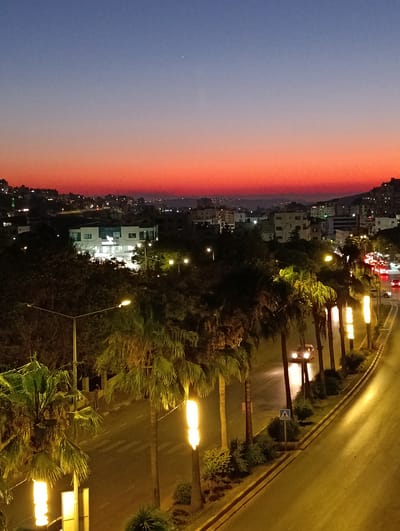 The image depicts a twilight scene in Nablus, Palestinian Territories, with a vibrant gradient of orange and purple in the sky. A city sprawls across rolling hills in the background, illuminated by the lights of buildings and streetlamps. In the foreground, a street lined with palm trees is visible. The street is wet, suggesting recent rain or mist, and glistens under the artificial lights. Several cars are on the road, with their headlights on, adding to the illuminated effect. Tall, vertical light poles with bright yellow lights are spaced along the street, casting a warm glow. Some of the palm trees appear to have lights near their bases, further enhancing the nighttime ambiance. The overall mood is serene and urban, capturing a moment of transition from day to night. There is a pedestrian crossing sign visible on the right side of the road.