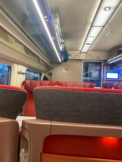 The image depicts the interior of a modern passenger train carriage, likely situated in or traveling through Saint-Denis, France. The perspective is from a low, seated vantage point, looking forward down an aisle toward the end of the carriage.

The immediate foreground features the backrests of two empty train seats, characterized by dark grey textured fabric with red piping along the top edge, resting on reddish-orange padded bases, all encased in light brown plastic shells. An orange light emanates from beneath the right foreground seat.

Overhead, the light beige ceiling is fitted with both continuous linear LED light strips and recessed circular spotlights, providing illumination. Along the left side of the carriage, transparent overhead luggage racks are visible, one containing a grey hard-shell suitcase. A dark blue neck pillow hangs from a fixed grab handle.

Large rectangular windows line the left wall, showing a blurred external environment and reflections of the interior. Towards the rear of the depicted compartment, a transparent glass door, featuring a map, leads to an adjacent section. To the right of this doorway, a glowing blue rectangular digital display is mounted on the wall. Various red and white informational or emergency signs are affixed to the beige wall panels. The carriage appears unoccupied in the visible area, with no clear individuals present.