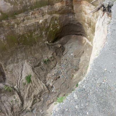 The image depicts a deep excavation or sinkhole in Kebunbaru, Indonesia. The scene is dominated by the raw, eroded earth walls of the pit, showing layers of sediment and rock. Patches of green moss and small plants are visible clinging to the damp surfaces, indicating a humid environment. The bottom of the pit is filled with rubble, soil, and a significant amount of scattered plastic debris, including blue and white fragments. A few branches and pieces of wood are also present, some embedded in the sides or resting near the top edge. To the right, a steep embankment of loose gravel and small stones leads down into the pit. The lighting suggests it is daytime, with no distinct shadows indicating a specific time. The overall impression is one of an untidy, possibly abandoned excavation site, affected by erosion and human waste. There is no visible text in the image.
