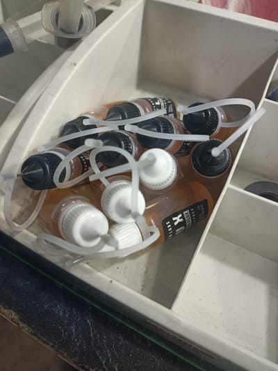 The image shows a white plastic organizer box containing several small bottles of e-liquid. The bottles are clear and contain an amber-colored liquid. Most of the bottles have black or white caps with thin, clear plastic rings around them. Some of the bottles have labels with text. One visible label reads "LUX FLUX" and below that "MADE IN INDONESIA". The organizer box appears to be in a well-lit indoor environment, possibly a workshop or a storage area. The arrangement of the bottles and the overall cleanliness of the organizer suggest that this is a collection of vaping supplies, likely for personal use or for sale. The image is taken from a slightly elevated angle, looking down into the organizer. The background is blurred, but appears to be a dark surface. There are no people visible in the image.
