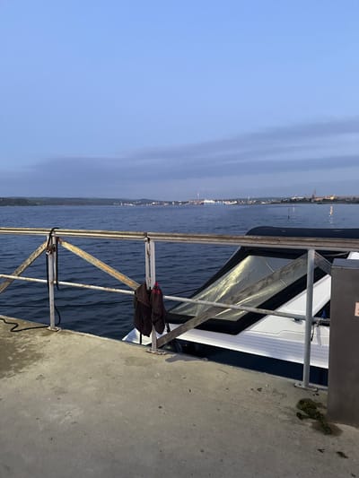 The image depicts a view of the Black Sea from a concrete pier in the city of Kazashko, Bulgaria. In the foreground, a white boat with a dark roof is docked. A metal railing runs along the edge of the pier, with two dark towels hanging from it. The water is calm, and in the background, a distant industrial shoreline is visible, characterized by cranes and smokestacks. The sky is overcast, with a pale blue hue, suggesting it is either early morning or late evening. The overall scene conveys a sense of quietude and industrial coastal activity.