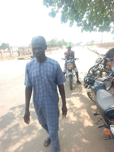 The scene is a daytime outdoor street environment in Nassarawan Eggon, Nigeria.
In the foreground, an adult male, approximately middle-aged, stands on a paved road, facing towards the camera. He wears a short-sleeved, blue and grey plaid tunic over matching trousers and dark flip-flops. Scarification marks are present on his left cheek. He holds a small, light-colored object in his right hand, positioned at his side. Overhanging tree foliage casts dappled shadows on the road surface around him.
In the mid-ground, to the right of the foreground figure, a younger male sits astride a light-colored motorcycle. He wears a light green beanie, a striped t-shirt (red, white, blue), dark trousers, and dark shoes. He is actively engaged with a mobile phone held in both hands. To his right, two additional motorcycles are parked; one is dark-colored with a black seat and visible amber turn signal, and another is partially obscured behind it.
The background features a paved road extending into the distance under a bright, clear sky. A low, light-colored concrete wall runs along the left side of the road, behind which are visible parts of buildings. A white sign with dark text, clearly displaying "Nassarawa Eggon," is affixed to the wall on the left. The overall environment appears dry and sunny.
