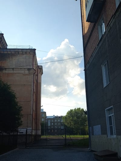 This vertically oriented image captures an outdoor scene on a bright, partly cloudy day, likely in the late afternoon given the angle of the sun. The perspective is from ground level, looking upwards and slightly between two buildings.

On the left, a tall, old, beige building with ornate architectural details is partially visible, showing part of its facade and the top of a fence. On the right, a dark, modern building with a brick and grey concrete exterior dominates the right side of the frame. It features multiple windows, some of which are white-framed. A dark balcony or overhang juts out from the brick section of this building.

Between the two buildings, the sky is a clear blue with a large, dramatic cumulus cloud formation. In the distance, beyond a black metal gate and a patch of green grass, is another residential building with a light-colored facade and many windows. A large, leafy green tree is prominent in the mid-ground, to the left of the distant building.

The scene is devoid of people, but several small, dark shapes, likely birds, are visible against the sky and clouds. Overhead power lines crisscross the sky, adding to the urban or suburban feel of the location. The ground in the foreground appears to be gravel. The overall impression is one of a typical residential area in a city. There is no visible text in the image.