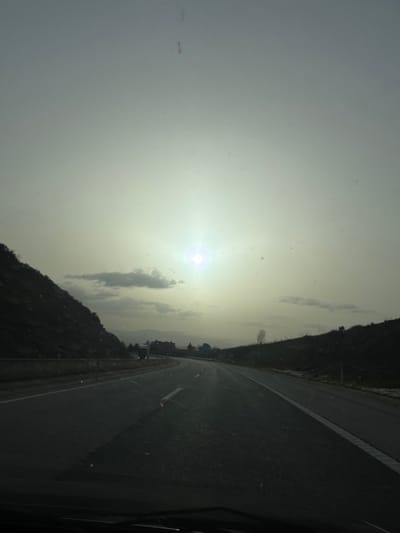 The image depicts a multi-lane highway scene, observed from inside a vehicle through a windshield marked with dust and debris. The highway extends into the distance, curving slightly to the left, with white lane markings visible on the dark asphalt surface. To the left of the road, a steep, dark, vegetated hillside rises, bordered by a concrete barrier or guardrail. To the right, a gentler, dark hillside also covered in sparse vegetation and some lighter patches, possibly snow or bare rock, parallels the road. A single leafless tree stands on the right hillside. In the mid-distance on the left lane, a dark-colored vehicle, possibly a truck, is visible. The sky is overcast and hazy, with a bright sunburst positioned low and slightly left of center, indicating the sun's location. Scattered dark clouds are present in the sky, particularly on the left. Distant mountains or hills are discernible on the horizon, with what appear to be structures of a small town or village nestled in a valley between them. This scene is located on a highway near Macedo de Cavaleiros, Portugal.