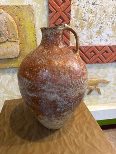 An ancient terracotta jug with a handle and a narrow neck sits on a wooden surface. The jug has horizontal lines around its body and is decorated with faded geometric patterns. The background features a wall with textured artwork, including a yellow relief and red geometric designs. A small, light-colored wooden sculpture of an animal is visible in the background, to the right of the jug. The scene is indoors, suggesting a museum or exhibition space in Fort-Şevtşenko, Kazakhstan.
