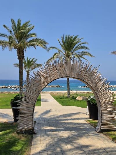 The photograph captures a sun-drenched outdoor scene in Hlorakas Community, Cyprus. In the foreground, a pathway paved with light-colored square stones extends into the distance. Flanking the pathway are patches of green grass. A prominent feature is a large, arch-shaped structure made of numerous thin, light-brown bamboo poles, arranged radially and fixed onto a curved frame. This bamboo arch serves as an entryway or decorative element. To the left of the arch, a large black planter holds a lush green plant.
Beyond the arch, the pathway continues towards a body of water, which appears to be the sea, with a clear blue hue. Along the shoreline, a line of dark rocky structures forms a breakwater. A small white boat is visible in the water near the shore. In the background, several palm trees with their distinctive fronds stand tall against a bright blue sky. The overall impression is a tropical or Mediterranean resort setting, likely a pathway leading to a beach or swimming area. The image is composed to highlight the bamboo arch and the inviting vista of the sea beyond.