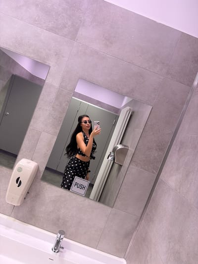 The image shows a woman taking a mirror selfie in a bathroom. The bathroom appears to be modern with gray tile walls and floor. In the reflection, there is a stainless steel hand dryer, a "PUSH" sign, and a sink with a chrome faucet. The woman is wearing a black and white polka dot outfit and sunglasses, holding her phone up to take the picture. The location is implied to be Orio al Serio, Italy, but there is no way to verify this using the visible elements of the picture.