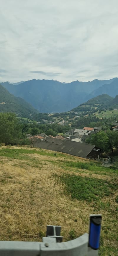 This media file is a landscape photograph taken in Challand-Saint-Victor, Italy. The scene depicts a village nestled on a mountainside, with rolling green hills and a backdrop of imposing blue mountains. The sky is overcast with fluffy white clouds, suggesting a mild or slightly cool day. The village itself is characterized by traditional stone houses with terracotta roofs, characteristic of Italian alpine architecture.
In the foreground, a portion of a grey metal railing with a blue grip is visible, suggesting the photo was taken from a vantage point, perhaps a balcony, observation deck, or even a vehicle. To the right of the railing, dry, golden grass covers the ground, interspersed with patches of vibrant green vegetation, indicating a change in season or landscape. The dry grass appears to be a field or a hillside.
The activity captured is a scenic view of the village and its natural surroundings. There is no specific event or human activity prominently featured, other than the implied presence of the photographer. The notable details include the clear layering of the mountains, the cluster of houses forming the village, and the contrast between the dry and green vegetation in the foreground. The time of day appears to be daytime, given the lighting, and the weather is likely overcast. No visible text is present in the image.