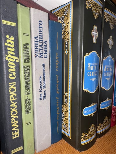 This media file shows a stack of books on a wooden surface, suggesting an indoor setting like a library or personal collection. The books appear to be old, with some featuring ornate designs on their covers, including gold and blue accents and religious imagery.
The visible text on the books is primarily in Russian and Belarusian. Some titles suggest collections of saints' lives and folk tales, with specific volumes marked by month ranges like "January-June" and "July-December." One book's title translates to "Street of the Younger Son," and another to "Russian-Belarusian Dictionary." The books are of varying colors, including black, blue, light gray, green, and dark gray.
The lighting suggests it is daytime, with a warm, natural light illuminating the scene. There are no people visible in the image, and no specific activity is depicted other than the display of the books. The overall impression is one of a well-read collection of literature, likely related to religious and cultural heritage. The location context provided, Chachersk, Belarus, reinforces the interpretation of the text and the subject matter of the books.