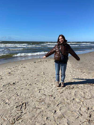A young woman stands on a sandy beach under a clear blue sky in Morskoye, Russia. She is captured in a mid-shot, looking directly at the camera with a gentle smile and her arms slightly outstretched. She has long dark hair and is dressed in a glossy dark brown puffer jacket, light blue jeans, and light brown sneakers with white laces. A black shoulder bag is worn across her chest.
The scene features a wide, light-colored sandy beach on the right, which meets a body of water with gentle waves on the left. The sky above is a vibrant, cloudless blue, indicating bright, sunny weather. The strong sunlight casts a distinct shadow from the woman towards the foreground, suggesting it is likely late morning or early afternoon. The sand beneath her feet is marked with numerous footprints and scattered with small pieces of driftwood or debris. The woman's relaxed posture and smile convey a sense of enjoyment of the outdoor environment. There is no visible text in the image.