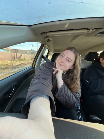 A woman is in a car. She is facing the camera, resting her head on her hand. She is wearing a brown jacket. There is another person in the back seat of the car. The car is moving past a street. It is a sunny day. The location is likely Yasenevo, Russia, based on visual features in the background such as building design and foliage.