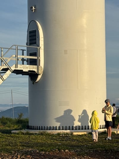 In Phát Chi, Vietnam, a group of individuals is gathered near the base of a large, white wind turbine. The turbine is cylindrical and features a metal staircase leading up to a small entrance with a slatted window. A light fixture is mounted near the top of the visible portion of the turbine.

Three children and two adults are visible in the foreground. One child, wearing a yellow hooded jacket and light-colored pants with striped cuffs, stands facing away from the camera, looking at the turbine. Another child, whose back is to the camera, is standing to the left of the first child, also facing the turbine. The third child is partially obscured behind the first two, and appears to be wearing a light-colored outfit.

An adult, wearing a light yellow collared shirt, dark shorts, a cap, and white sneakers, stands to the right of the children, facing them. This adult appears to be holding a small object. Another adult, wearing dark clothing, is visible further to the right, behind the first adult.

The ground around the turbine is covered in green grass and some scattered rocks and dirt. In the background, a hazy landscape of rolling hills and mountains can be seen under a blue sky with some clouds. Power lines are also visible in the distance. The shadows of the children and the turbine are cast on the side of the turbine.