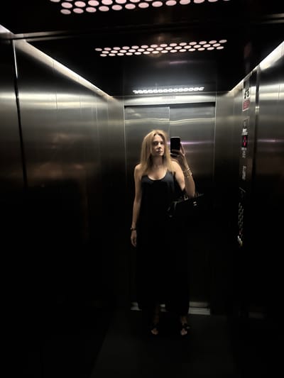 A woman is taking a selfie in an elevator. She is wearing a black tank top and several gold bracelets on her left wrist. She is holding her phone in her right hand to take the picture. The elevator has stainless steel walls and the floor appears to be made of the same material. The elevator also has a panel of buttons on the wall. The lighting in the elevator is dim, with some lights on the left wall that appear to be part of the elevator's design. The image is taken from a slightly low angle, looking up at the woman. The overall mood is casual and private. There is no visible text in the image. The location context is Loures, Portugal.