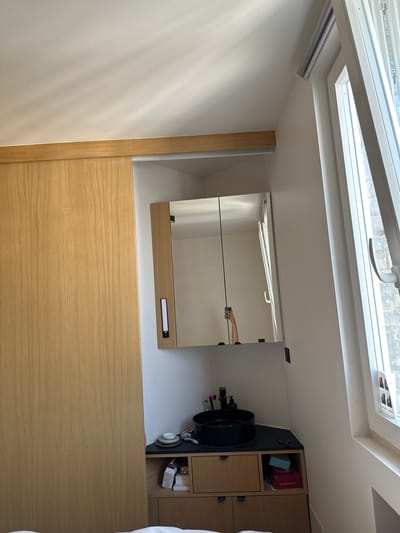 This appears to be an interior shot of a room, possibly a bathroom, with a modern aesthetic. The location is Saint-Ouen-sur-Seine, France.
The image is taken at an angle, showing a portion of the ceiling with a skylight. The skylight frame is white, and a window is visible.
Below the skylight, there is a white wall that connects to a wooden paneled wall. There is a mirror mounted on the wall. A reflection of a hand can be seen in the mirror.
Adjacent to the mirror and wooden paneling, a cabinet with drawers and shelves can be seen. There are items stored on the shelves and a black sink is to the left of the cabinets.