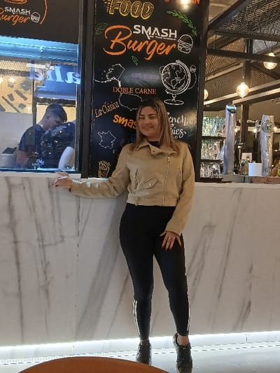 A woman in a tan jacket and black leggings poses in front of a "Smash Burger" restaurant sign. The sign features burger and globe illustrations with menu items. She is standing on a white marble floor, with a reflection visible beneath the sign. A man is seen in the reflection, partially obscured. The restaurant's interior includes metal beams and various food service equipment.