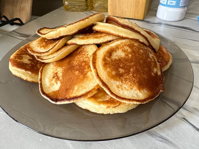 This is a close-up, overhead shot of a stack of golden-brown pancakes on a dark grey plate. The pancakes are varied in size and shape, with some appearing thicker and fluffier while others are thinner and flatter. Their surfaces show a beautiful gradient of cooked dough, ranging from pale yellow to rich, toasted brown, with some slightly darker, crispier edges. The plate itself has a subtle, swirling pattern and rests on a light-colored, textured surface that appears to be a countertop with a marble-like design.
In the background, out of focus, are elements that provide some context. To the left, a wooden object, possibly a cutting board or a decorative item, is visible. Behind the pancakes and to the right, a partially obscured white plastic container with blue lettering is present. The visible text on the container includes "ГОСТ" and "500 г", suggesting it may contain a food product measured in grams, compliant with Russian state standards. A blurred bottle of cooking oil can also be seen.
The lighting suggests it is daytime, possibly in a kitchen or dining area. The overall impression is that of a homemade breakfast or snack, with the focus entirely on the appetizing presentation of the pancakes. There are no people visible, and the scene evokes a feeling of domestic comfort and simple culinary pleasure. The location context of Засечное, Russia, aligns with the presence of Russian text on the container.