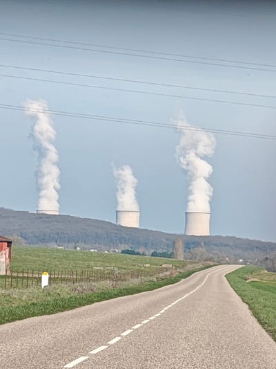 The image depicts a rural scene leading towards the Cattenom Nuclear Power Plant, located near Thionville, France. In the foreground, a two-lane asphalt road with a dashed white center line curves gently to the right as it recedes into the distance. Green grass verges border both sides of the road. On the immediate left, a partial view of a red-sided building or shed is visible behind a wire fence. A white marker post with a yellow top stands near the fence line on the left. The midground features a gently sloping green field, transitioning to a low hill densely covered with bare deciduous trees. Isolated residential structures are faintly visible within the wooded area. In the background, three large, light-colored, hyperbolic cooling towers of a nuclear power plant rise above the tree line. Each tower emits a voluminous, turbulent white plume of steam or water vapor that ascends vertically against a clear, pale blue sky. Multiple thin, dark power lines horizontally traverse the upper portion of the frame, intersecting the rising plumes. No people or vehicles are present.
