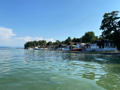 The image depicts a serene lakeside scene in Milvignes, Switzerland, likely at a recreational area. The foreground is dominated by the clear, tranquil waters of a lake, showing subtle ripples and reflecting the bright blue sky and surrounding foliage. In the midground, a line of small, modern structures with dark-tiled roofs runs along the waterfront. These structures appear to be cabins or boathouses, some with white awnings or umbrellas deployed. Several boats are moored at a wooden pier in front of these buildings, including a small red boat, a sailboat covered with a tarp, and a white motorboat. A few people are visible in or near the water; one person is standing in the water near the pier, and others are on the pier or the shore. The background features a dense line of green trees and vegetation, with distant mountains faintly visible on the horizon under a clear blue sky. The overall impression is one of a peaceful summer day at a lakeside resort.