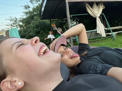 Two young women are lying on their backs outdoors. The woman in the foreground is laughing with her mouth open wide, showing her teeth. She is wearing a black t-shirt and a silver stud earring in her left ear. The woman behind her is wearing a black t-shirt and a pink baseball cap. She is sticking her tongue out at the camera and smiling. Both women appear to be relaxed and enjoying themselves.
In the background, there are trees, a clothesline with laundry hanging on it, a covered swing set, and another woman sitting in a green chair, holding a colorful pinwheel. The scene appears to be a backyard on a sunny day. The time of day seems to be late afternoon or early evening, as the lighting is soft and warm. The overall mood is lighthearted and joyful. There is no visible text in the image.