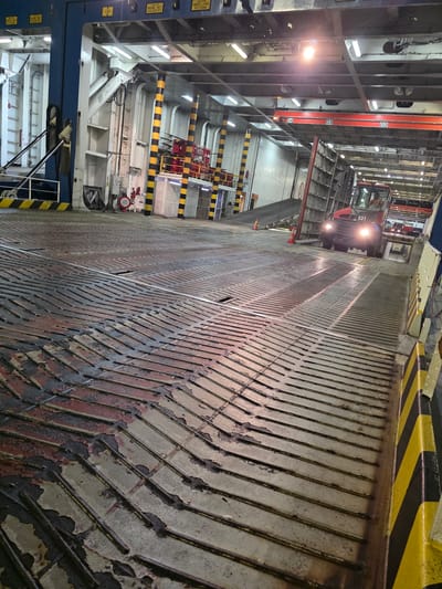 The image shows the interior of a ferry's car deck. The deck is made of metal with a patterned surface, showing wear and tear with some reddish staining. In the background, a small red vehicle, possibly a tugger or a small forklift, is visible with its headlights on. The vehicle has the identification "S31" on its front. Other structures of the ferry, including support beams, railings, and lighting, are visible. There are yellow and black striped markings on the floor and walls, indicating safety zones. The scene appears to be in a working industrial environment, likely within a port facility. No specific details about the city of Ravenna, Italy are visible in the image itself.