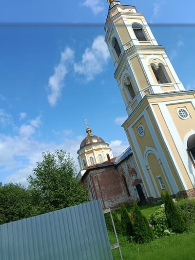 The image shows a bell tower and a church dome against a blue sky with white clouds. A corrugated metal fence runs along the bottom of the frame, and there are trees and manicured bushes in the foreground. The bell tower is painted yellow with white trim and has arched openings. The church dome is golden and topped with a cross. The church building itself is made of red brick with white architectural details. The scene appears to be located in the village of Pashnevo, Russia, judging by the architectural style and the overall setting. There are no people visible in the image.