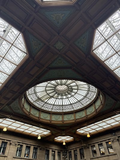 This is a low-angle, full shot of the interior of the Scottish National Gallery in Edinburgh, United Kingdom, looking up at the ornate ceiling and a domed skylight. The ceiling is a complex structure of dark wood panels with decorative teal and gold friezes. The central focus is a large, circular skylight with a radiating pattern of glass panes and metalwork, surrounded by a decorative band of figures and foliage. Flanking the central dome are smaller, rectangular skylights, also featuring intricate metalwork. Below the skylights, the walls of the gallery are visible, adorned with classical architectural details and rows of windows. The overall impression is one of grandeur and historical architecture.