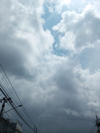 The image captures a daytime scene in Vieng Phouka, Laos, primarily focused on an overcast sky and urban infrastructure. The upper right quadrant of the sky displays patches of clear blue, with visible sunrays emanating from behind brighter clouds. The remainder of the sky is filled with a large formation of cumulus and cumulonimbus clouds, ranging in color from bright white, where illuminated by the sun, to dark grey, indicating substantial cloud cover. In the lower left, a utility pole with multiple cross arms supports a dense, tangled network of black electrical and communication wires that traverse horizontally across the lower portion of the frame. A street lamp extends from this pole. The tops of building structures with light-colored facades are partially visible beneath the wire infrastructure in the bottom left corner. No individuals or specific actions are depicted.
