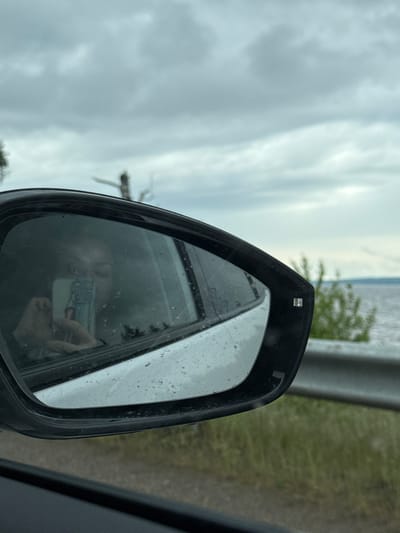 The image, rotated 90 degrees counter-clockwise, provides a view from inside a car, focusing on its side mirror. The mirror's surface is covered with small water droplets, indicating recent rain or a damp environment.

Within the reflection of the mirror, a person's hand is clearly visible, holding a light-colored smartphone, which is presumably being used to take the photograph. A portion of the person's face is also faintly visible in the reflection. A small, non-textual symbol, possibly an indicator for a blind spot monitoring system, is discernible on the mirror's surface.

The external environment seen outside and around the mirror consists of a predominantly cloudy or overcast sky. To the right of the mirror, blurred green foliage and a grey vertical structure, likely a roadside barrier or fence, suggest a location alongside a road. The immediate foreground on the right shows a part of the car's dark interior, such as a door panel. A small, clear rectangular element is present on the top edge of the mirror housing, possibly a sensor or light.

The scene captures a moment of travel or a stop in Kryukovo, Russia, characterized by cloudy weather and damp conditions.