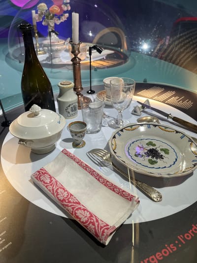 This image displays a historical dining table setting presented as an exhibit within the Cité de la Gastronomie et du Vin in Dijon, France. The objects are arranged on a circular white platform, partially enclosed by a clear, reflective acrylic dome.
In the foreground, a folded white linen napkin features a prominent red and white intricate pattern along its border. To its left, a white ceramic lidded soup tureen with decorative handles and faint blue floral motifs rests. Behind the tureen, a dark green glass bottle stands upright. A small, multi-colored ceramic egg cup with floral designs is positioned between the napkin and the tureen.
Further back in the display, a plain white stoneware jar, partially labeled "GREY," is placed near a tall brass candlestick holding a lit white candle. To the right, a clear glass tumbler and a stemmed wine glass are arranged. An oval-shaped ceramic plate dominates the right side, showcasing a hand-painted central motif of dark grapes and green leaves, encircled by blue and yellow decorative bands. A silver fork and a knife with a wooden handle are positioned next to the plate, accompanied by an additional silver spoon and a small white triangular condiment holder.
The background features abstract blue and purple ambient lighting, creating reflections on the dome. Another, smaller table setting is discernible further back within the enclosed space. Partially visible French text on the dark perimeter surrounding the white display platform indicates informational labeling. The scene is devoid of people.