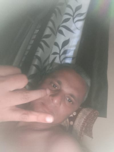 A man with short, graying hair is lying on a pillow, facing the camera. He is making a hand gesture with his index and pinky fingers extended. A curtain with a leaf pattern is visible behind him. The image is blurry and partially obscured by a light haze. The location is Dhandhuka, India, but no further contextual information is provided about the city.