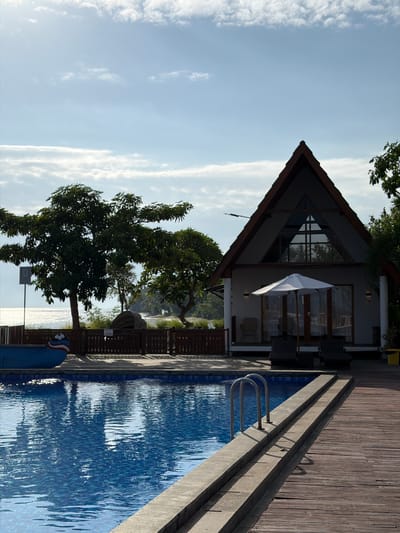 A resort setting is depicted in Pasir Putih, Indonesia, featuring a swimming pool and a triangular-roofed building. The pool, filled with blue water, has a tiled bottom and is bordered by a concrete ledge and a wooden deck. A metal ladder with two arched handrails provides access to the pool. In the foreground, a portion of a blue boat with red and white stripes is visible on the left. Behind the pool, a wooden fence separates the area from the building and surrounding vegetation.
The building, painted white, has a prominent triangular roof with dark brown trim. A large triangular window is situated at the peak of the roof. In front of the building, a white umbrella shades two dark lounge chairs. Two additional chairs and a small table are visible on the covered porch.
To the left of the building, large trees with dense green foliage are present, with the shimmering ocean and a distant shoreline visible in the background. The sky is a bright blue with scattered white clouds. The overall scene suggests a serene tropical environment.