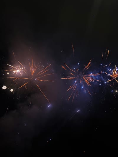 The image shows a nighttime scene with fireworks illuminating a dark sky. Several bursts of fireworks are visible, with streaks of orange, yellow, blue, and white light spreading outwards. Wisps of smoke are also present, adding to the atmospheric effect. The setting appears to be outdoors, likely a public space where fireworks displays are held. There are no people or specific city landmarks clearly identifiable in the image, making it impossible to definitively confirm a location in Tutrakan, Bulgaria.