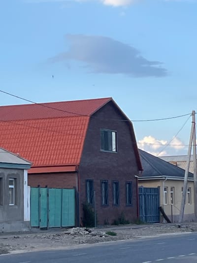 The image captures a view from a roadside in Balykchy, Kyrgyzstan. The scene is dominated by the facades of several buildings, suggesting an urban or semi-urban environment. In the foreground, a building with a striking orange-tiled roof is visible. Adjacent to it is a dark-colored building with a pitched roof and several horizontal windows. Further in the background, a lighter-colored building with multiple windows is partially obscured by the darker structures.
A prominent feature is a series of horizontally stacked, light blue structures, possibly shipping containers or modular units, positioned against the side of one of the buildings. A paved road runs alongside the buildings, with a distinct white lane marker visible. The sky above is a clear blue with some wispy clouds, and a single, small airplane can be seen in the distance. A utility pole with wires extends across the upper portion of the frame. The overall impression is one of a slightly overcast day, possibly late afternoon or early evening, given the quality of light. There is no discernible activity or human presence in the image, focusing solely on the architectural elements and the surrounding environment. No text is visible in the image.